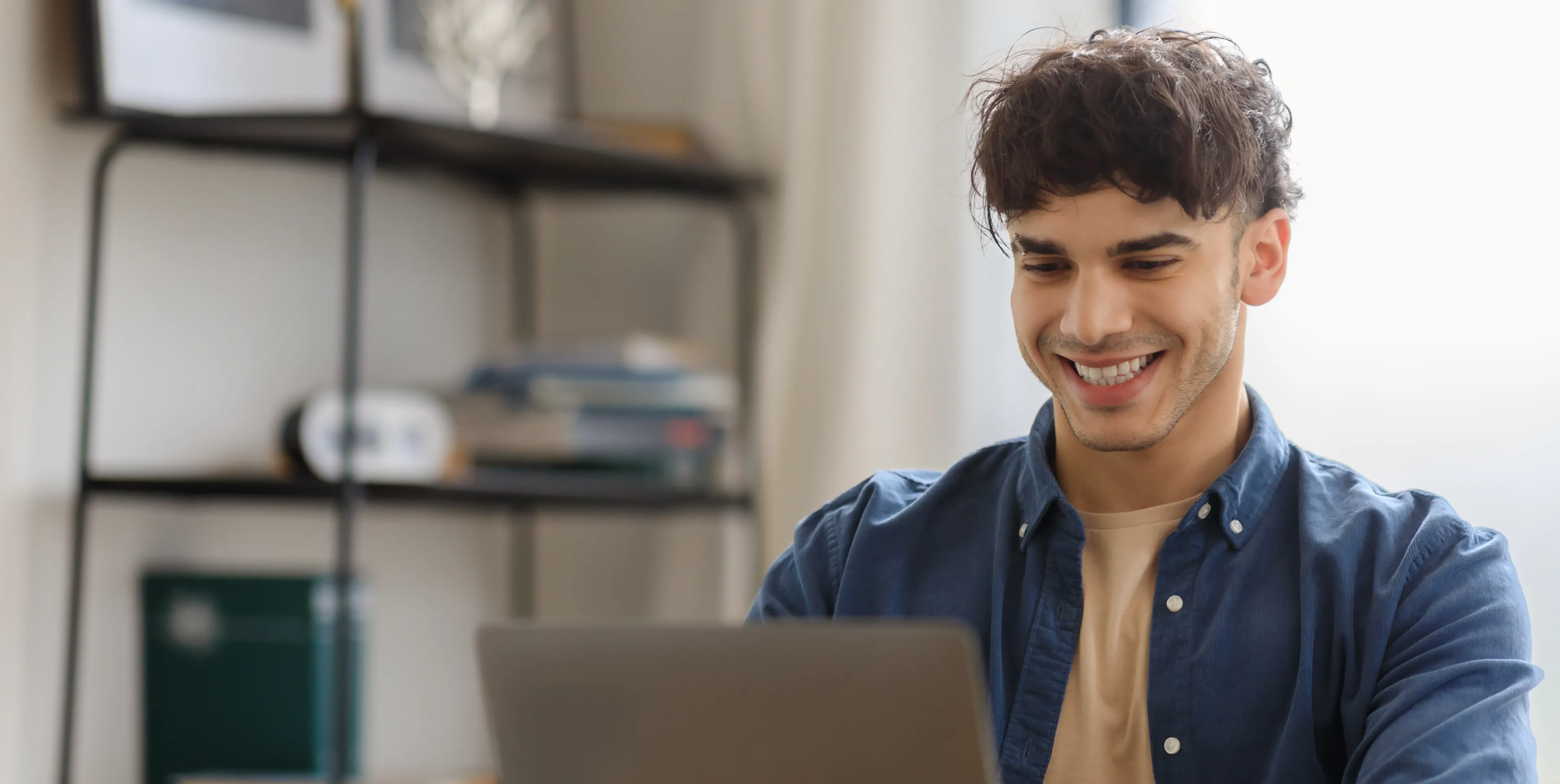 Smiling man sitting at a table, looking at his laptop screen in a bright room.