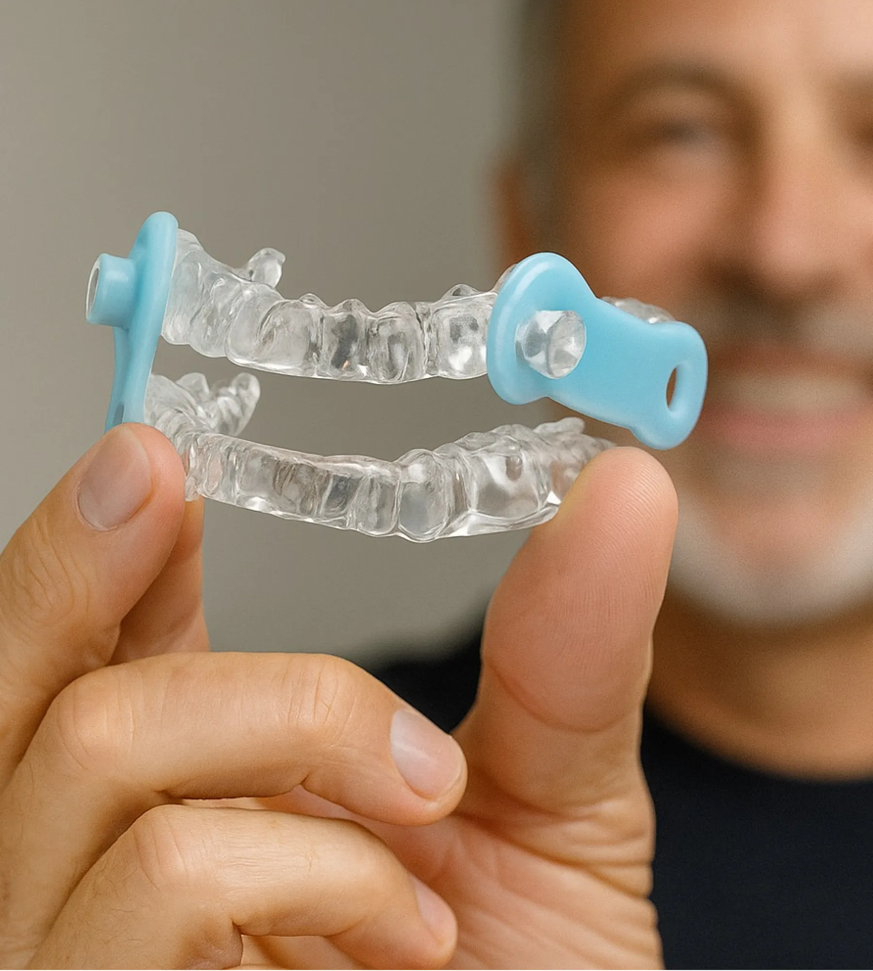 Person holding a clear dental mouthguard with blue ends, smiling in the background.