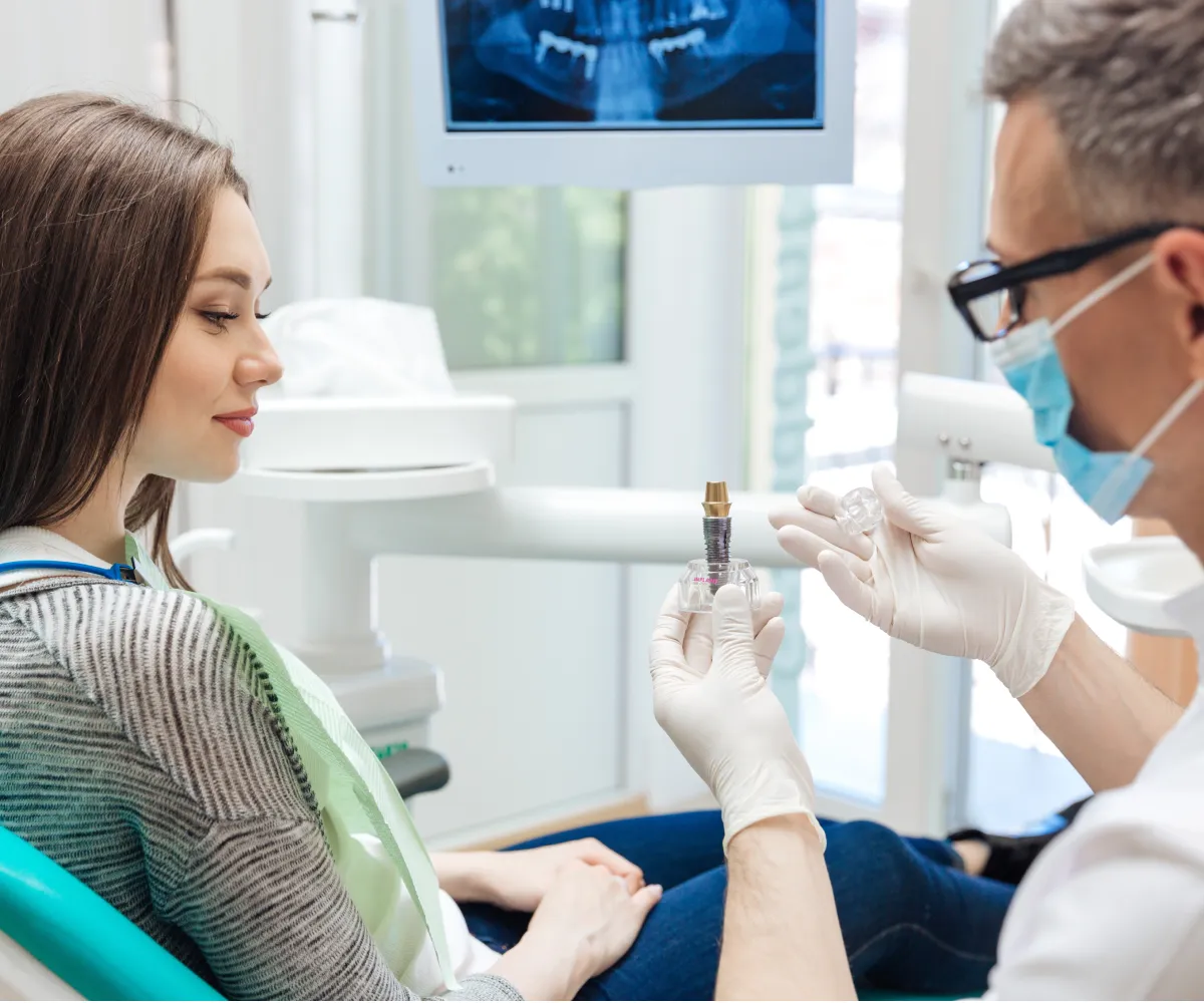 A dentist shows a dental implant model to a patient sitting in a dental chair.