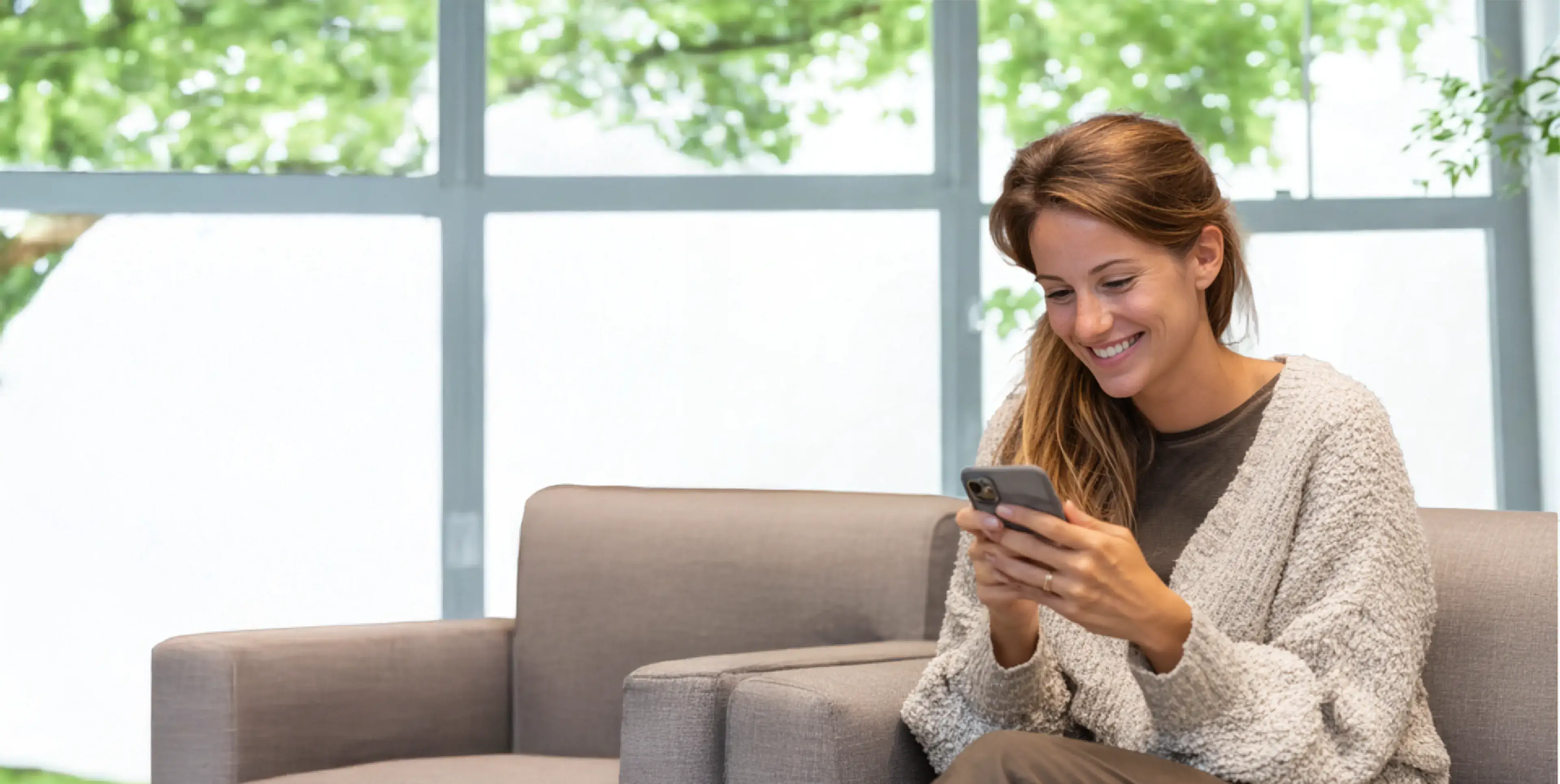 A woman sitting on a sofa, smiling while looking at her phone.