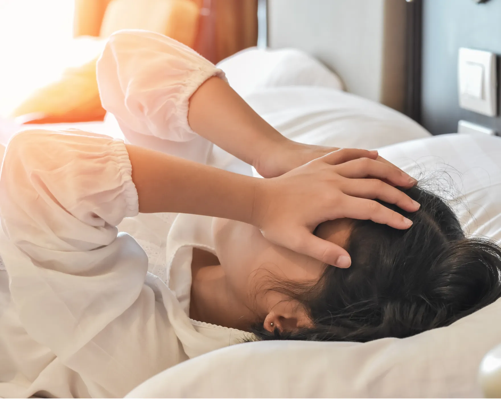 Person lying in bed, holding their head with both hands, appearing distressed.