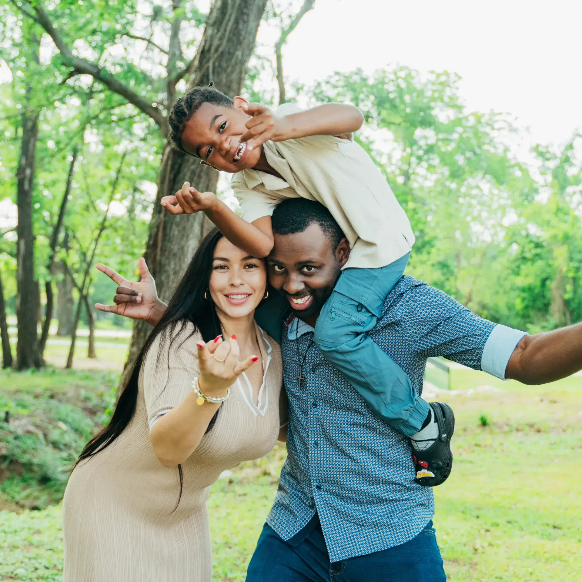 A man holds a smiling boy on his shoulders while a woman stands beside them in a park.
