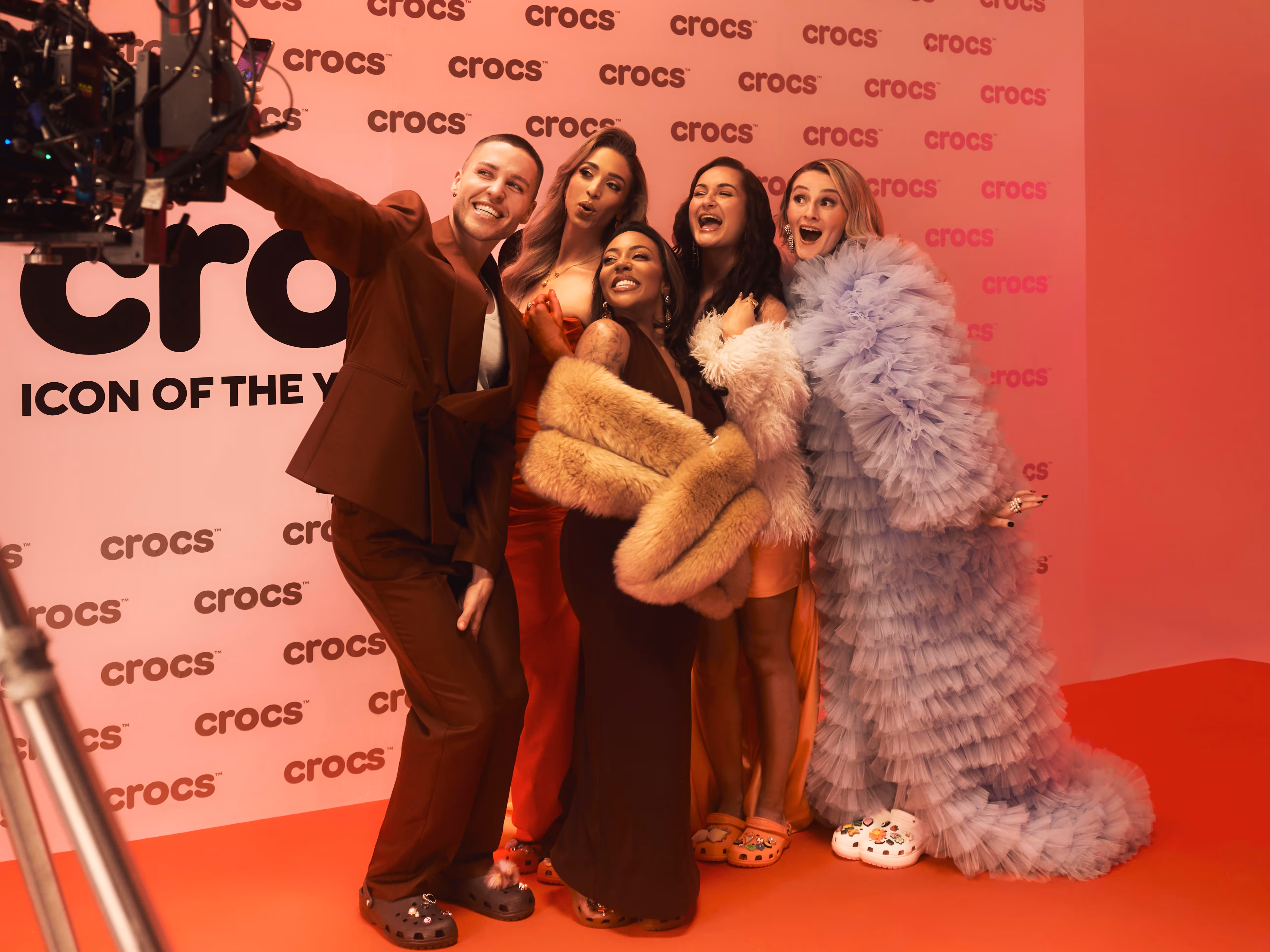 Crew operates camera equipment filming a woman in a red dress on a red carpet with Crocs branding.