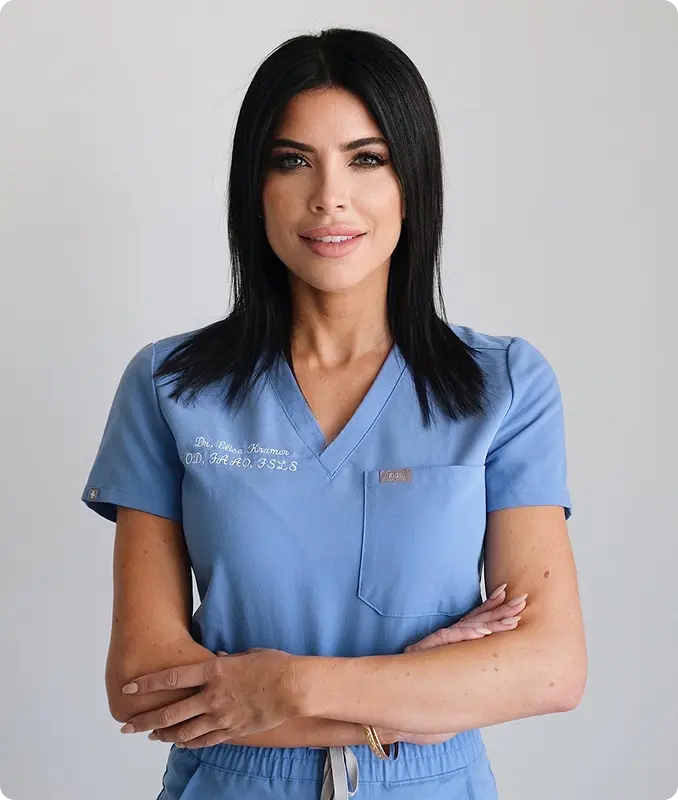 Smiling woman with black hair in blue medical scrubs with arms crossed against a gray background.