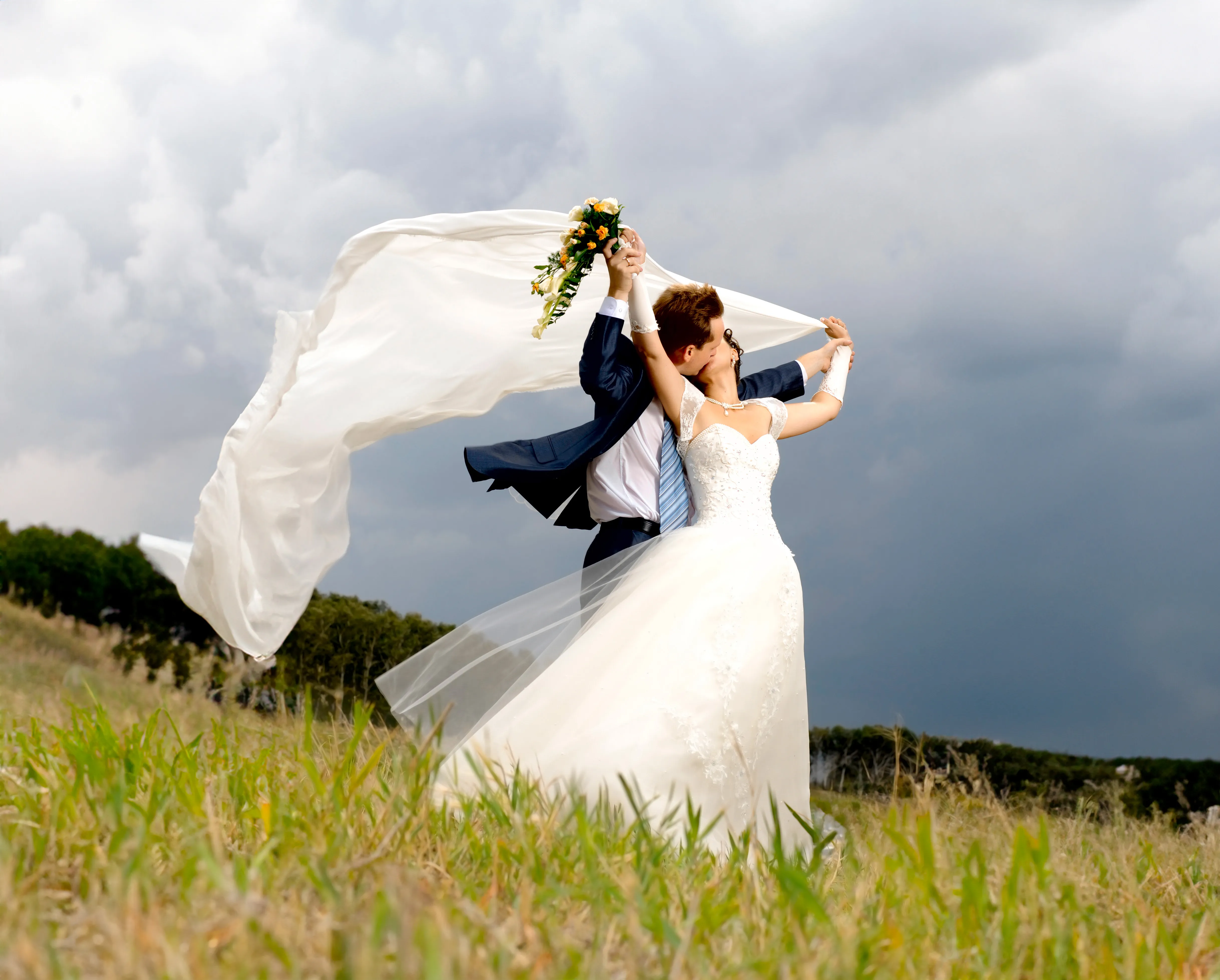 A bride and groom stand outdoors in a grassy field, holding a bouquet and lifting the bride’s veil as it flows in the wind under a cloudy sky.