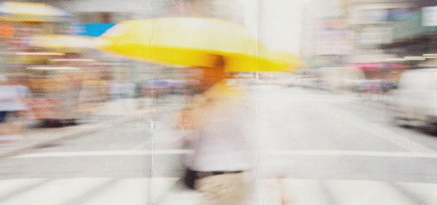 Person wearing light-colored clothing walking across a city street holding a bright yellow umbrella on a rainy day, with blurred urban background.