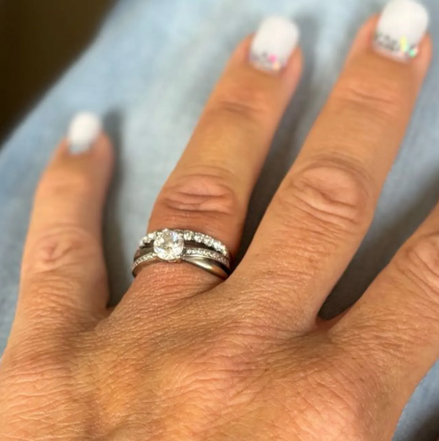 Main avec une bague en diamant et des alliances sur l'annulaire, ongles manucurés blancs avec des paillettes.