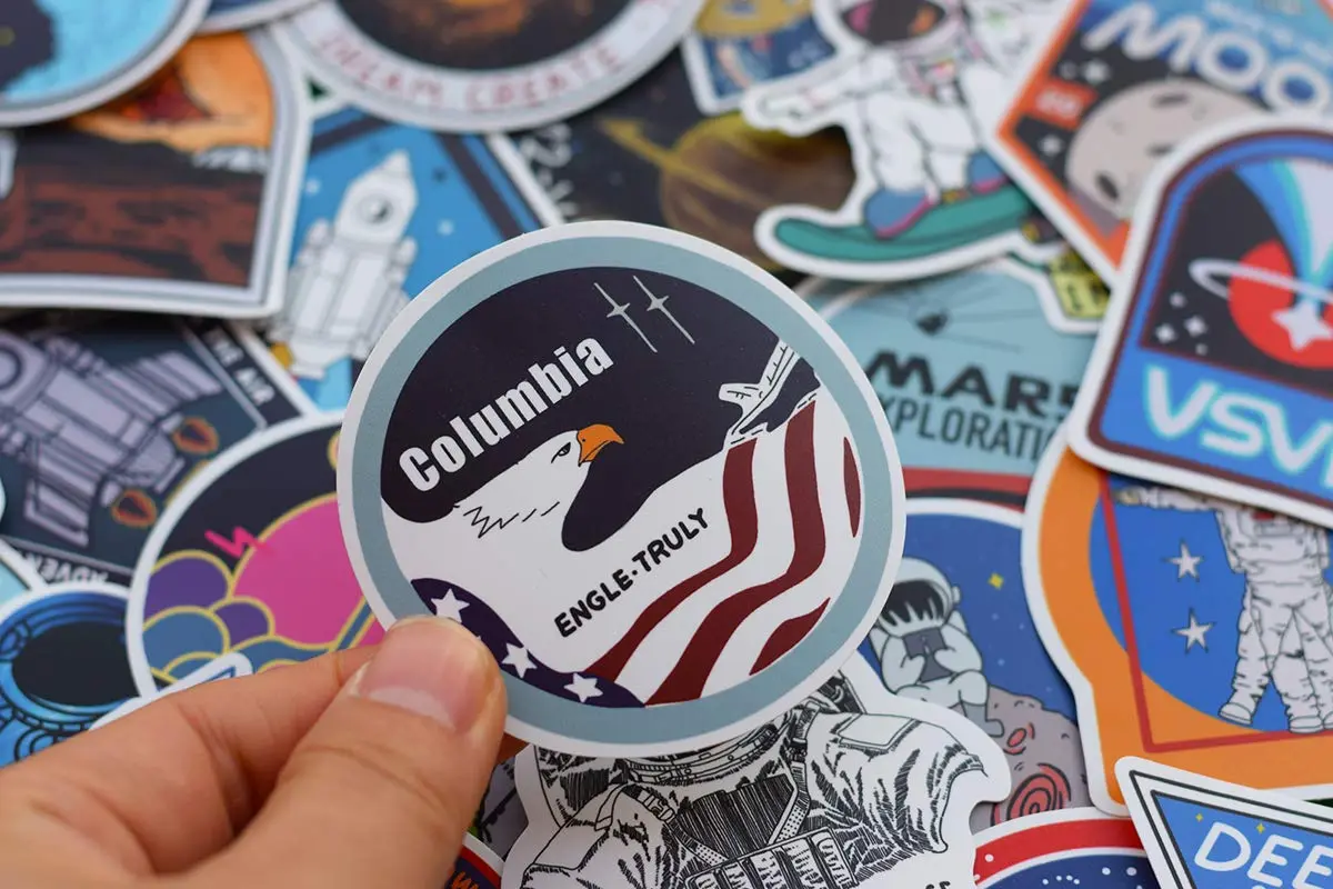 Autocollant de la navette spatiale Columbia et l'aigle américain tenu par une main, entouré d'une collection d'autres autocollants sur le thème de l'espace et de la NASA.