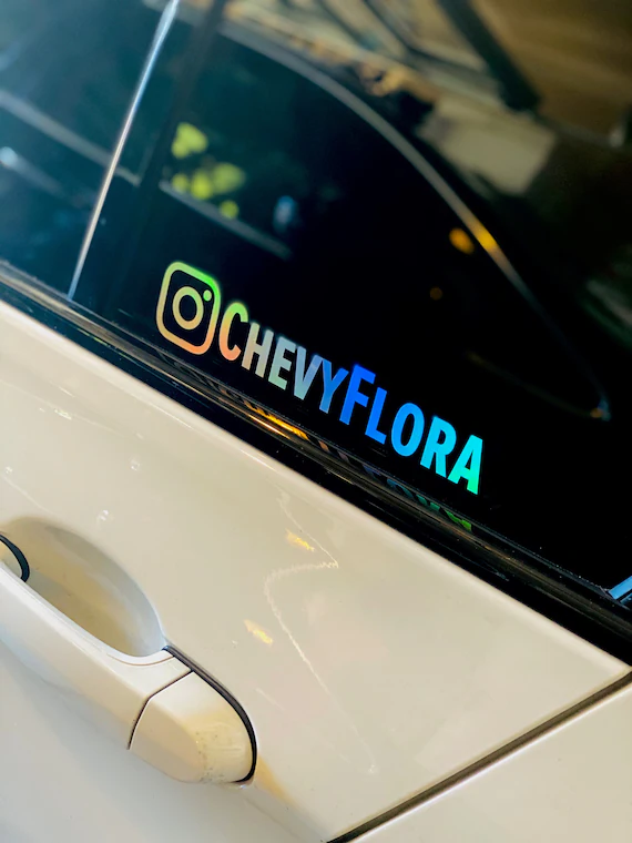 Sticker avec le logo Instagram suivi d'un pseudo en dégradé bleu sur la vitre arrière d'une voiture blanche.