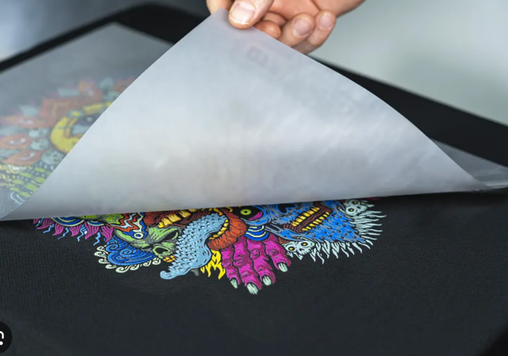Main soulevant un papier transfert thermique pour révéler un motif coloré complexe sur un tissu noir.