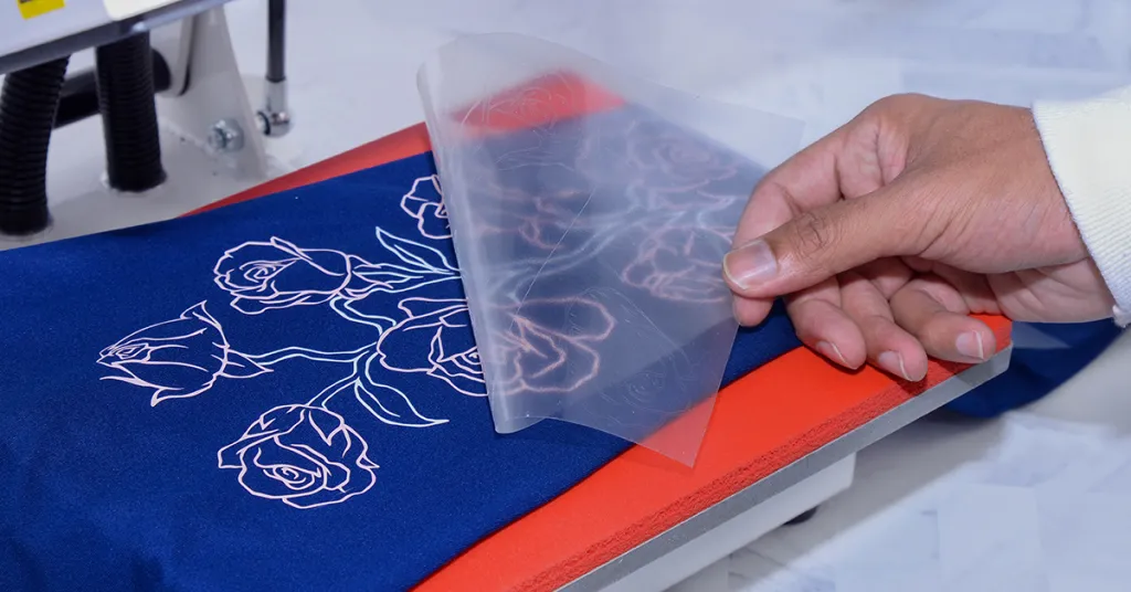 Main retirant un papier transfert collant d'un flocage sur un tissu bleu avec un motif de roses blanches.