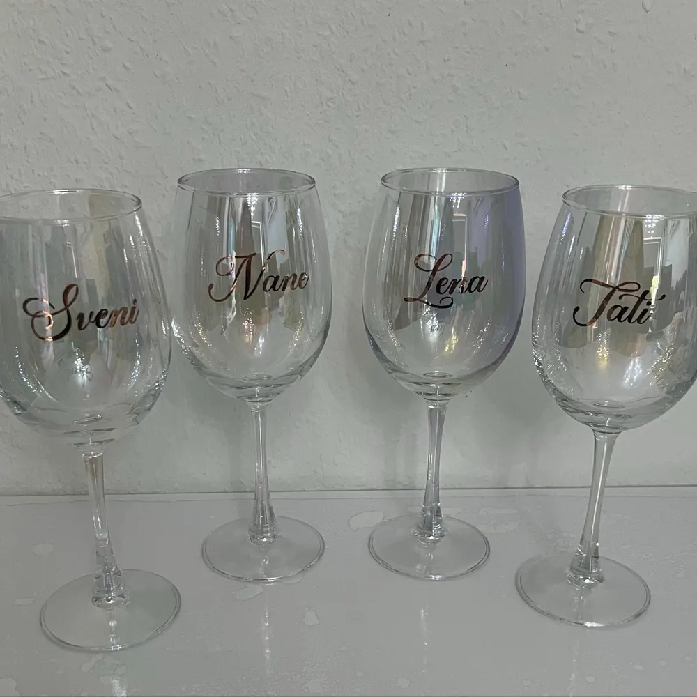 Quatre verres à vin transparents alignés, chacun avec un nom écrit en lettres cursives : Sveni, Nane, Lena, Tati.
