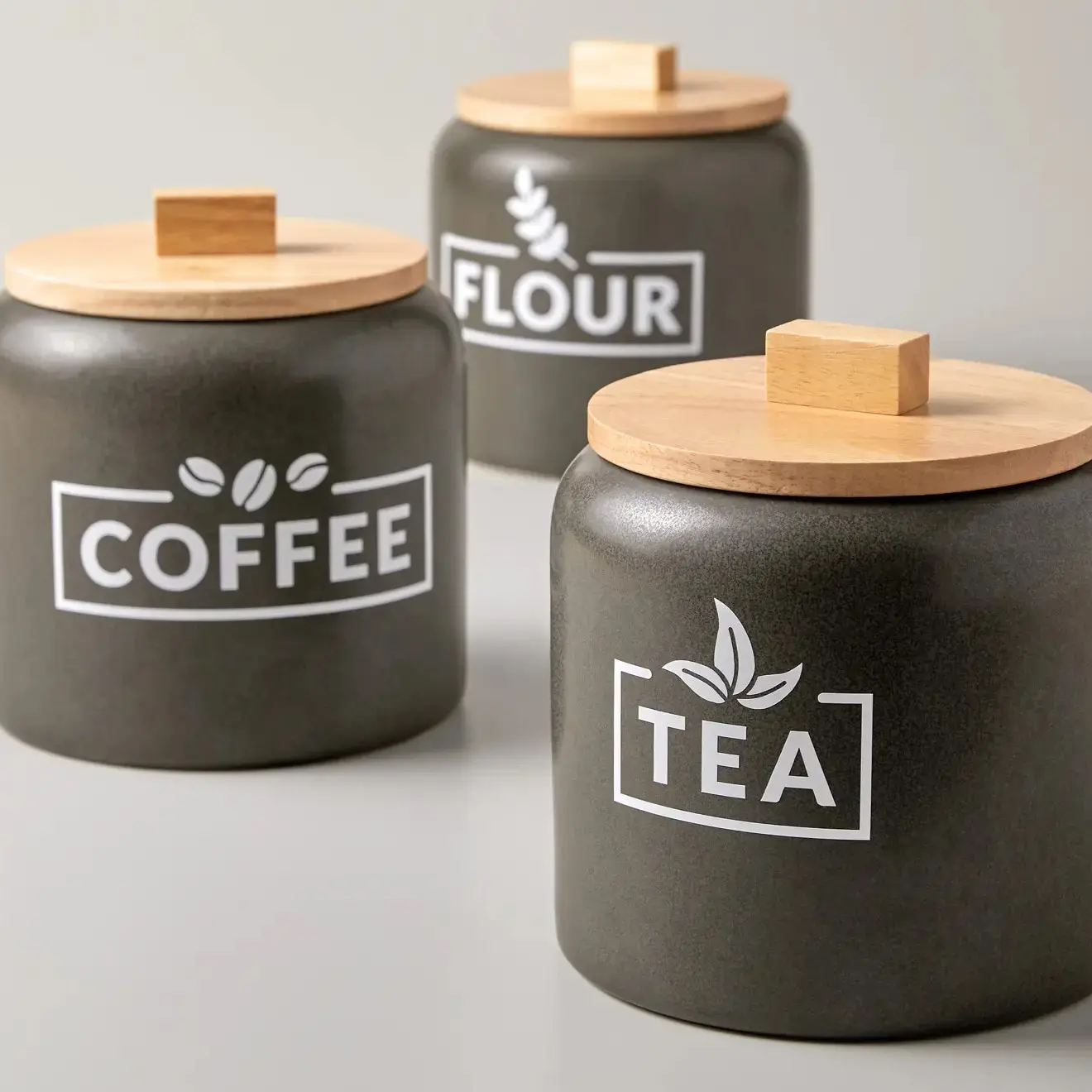 Trois pots de stockage noirs mats avec couvercles en bois clair, étiquetés "THÉ", "CAFÉ" et "FARINE".