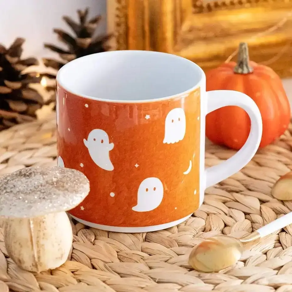 Tasse orange décorée de petits fantômes blancs posée sur une nappe en fibres naturelles avec une petite citrouille, un champignon décoratif, deux cuillères blanches et dorées, et un pomme de pin en arrière-plan.