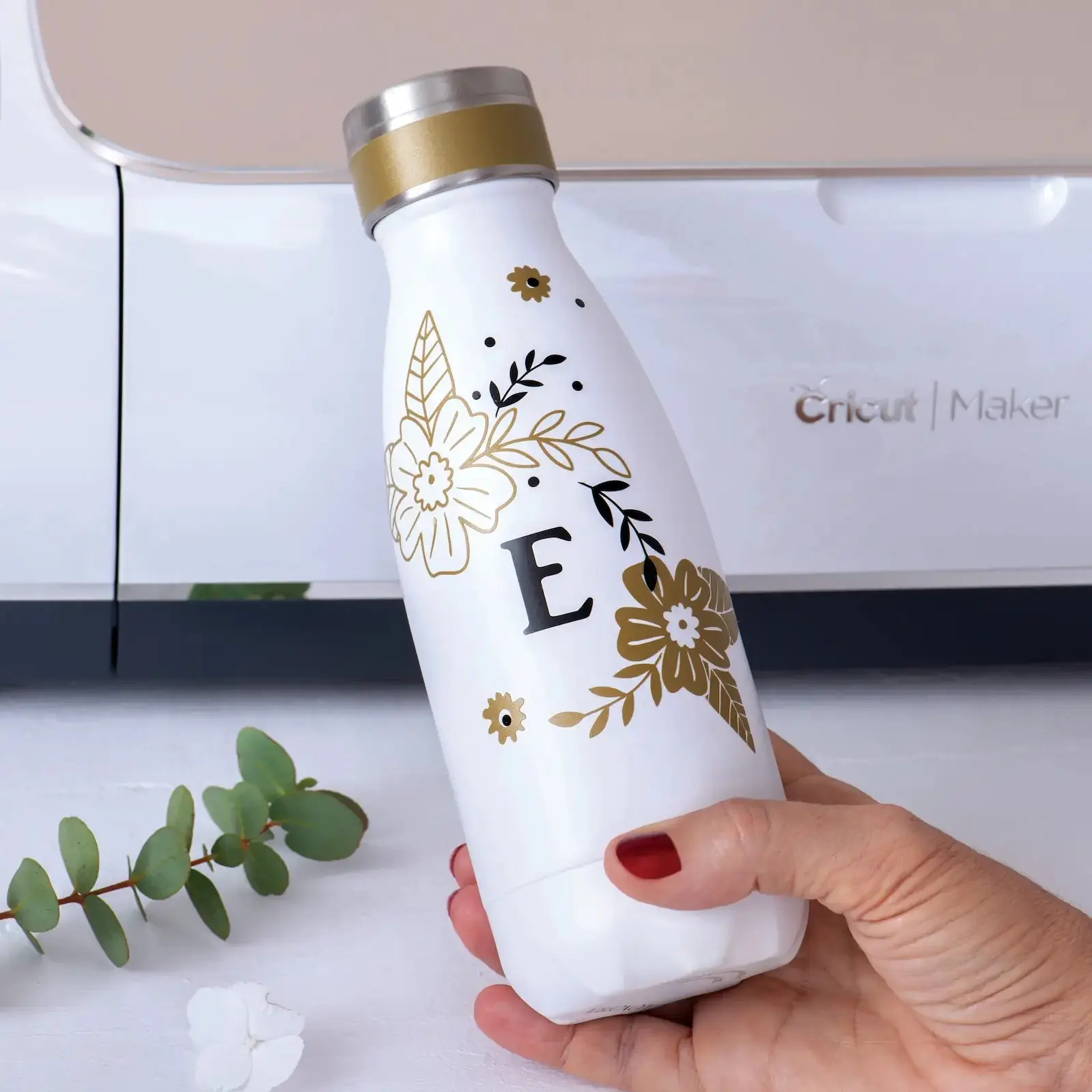 Une gourde blanche ornée de fleurs dorées et d'une lettre E noire tenue par une main, avec un appareil Cricut Maker en arrière-plan.