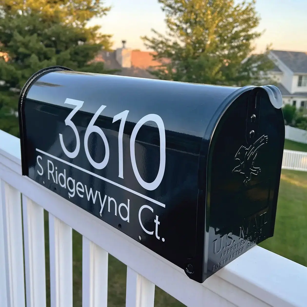 Boîte aux lettres noire avec une adresse imprimé dessus, fixée sur une rampe blanche, avec une pelouse et des maisons en arrière-plan.