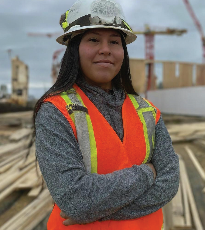 Female construction worker wearing an orange safety vest and white hard hat. She is standing with her arms crossed, looking straight at the camera.  