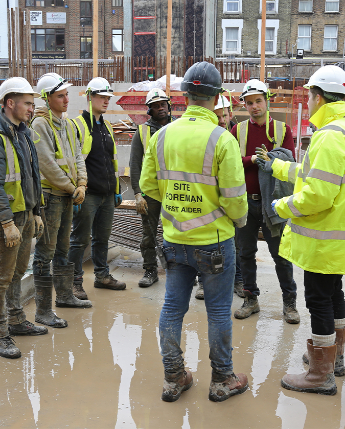 Eight construction working standing in a circle talking with each other. Around them is construction equipment and the ground is very wet.    