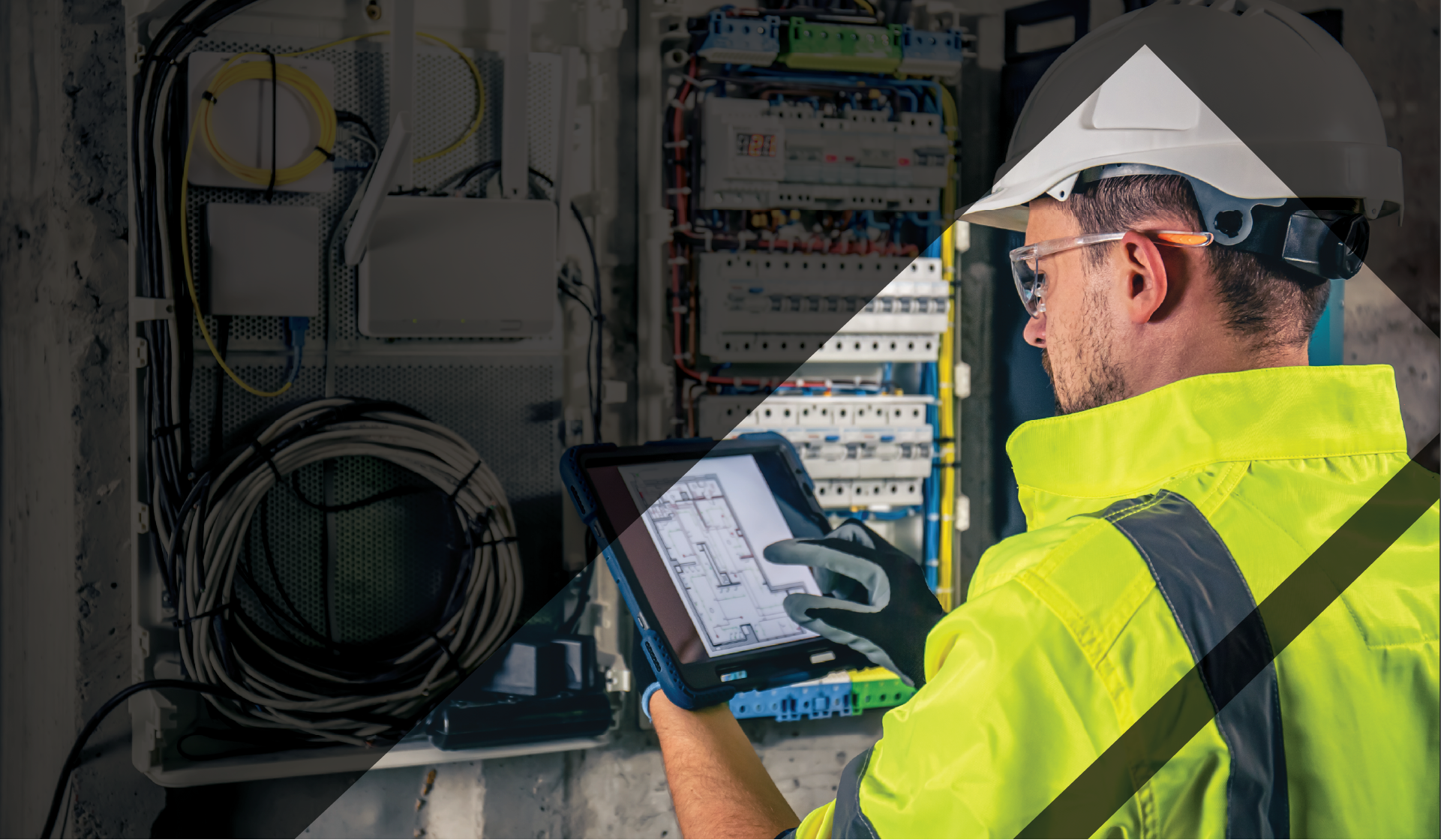 A construction working holding an Ipad looking at Blueprints. Around them is electrical wires and boxes.  