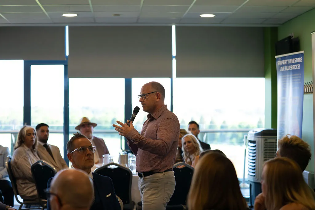 A man talking into a microphone surrounded by an audience at a property networking event
