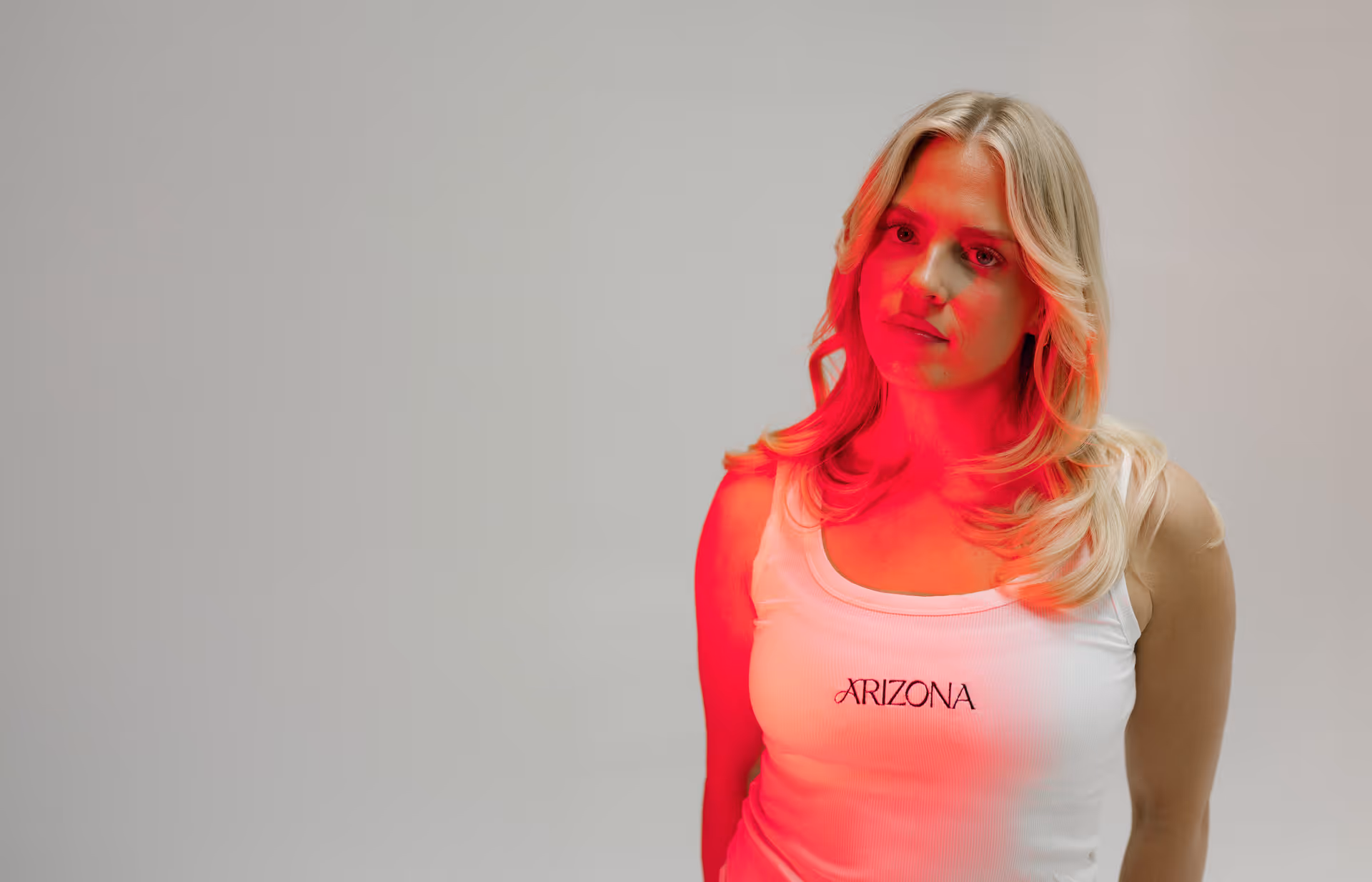 Woman in an Arizona-branded white tank top bathed in red light, looking directly at the camera against a neutral grey background.