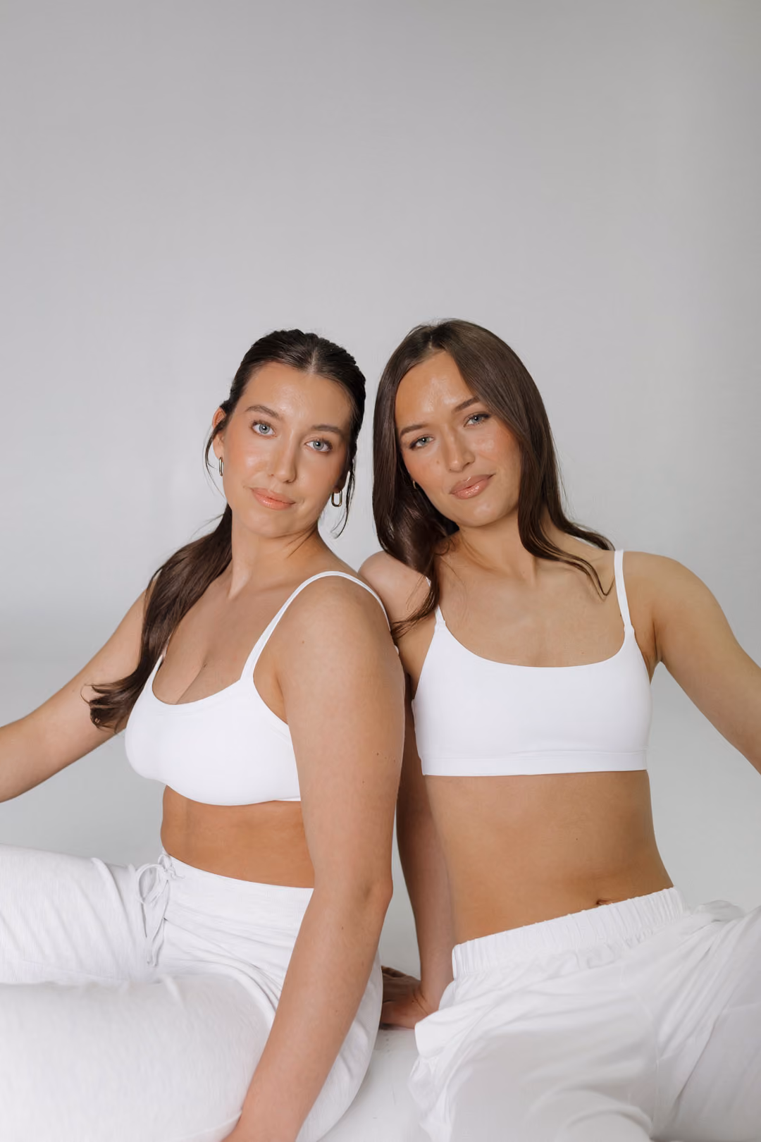 Two women in white tops and pants sitting close together against a light gray background.