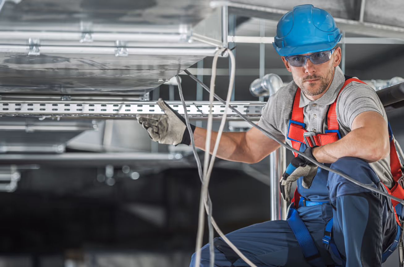 Commercial HVAC installer in a blue hard hat and harness working on overhead ductwork inside a large building.