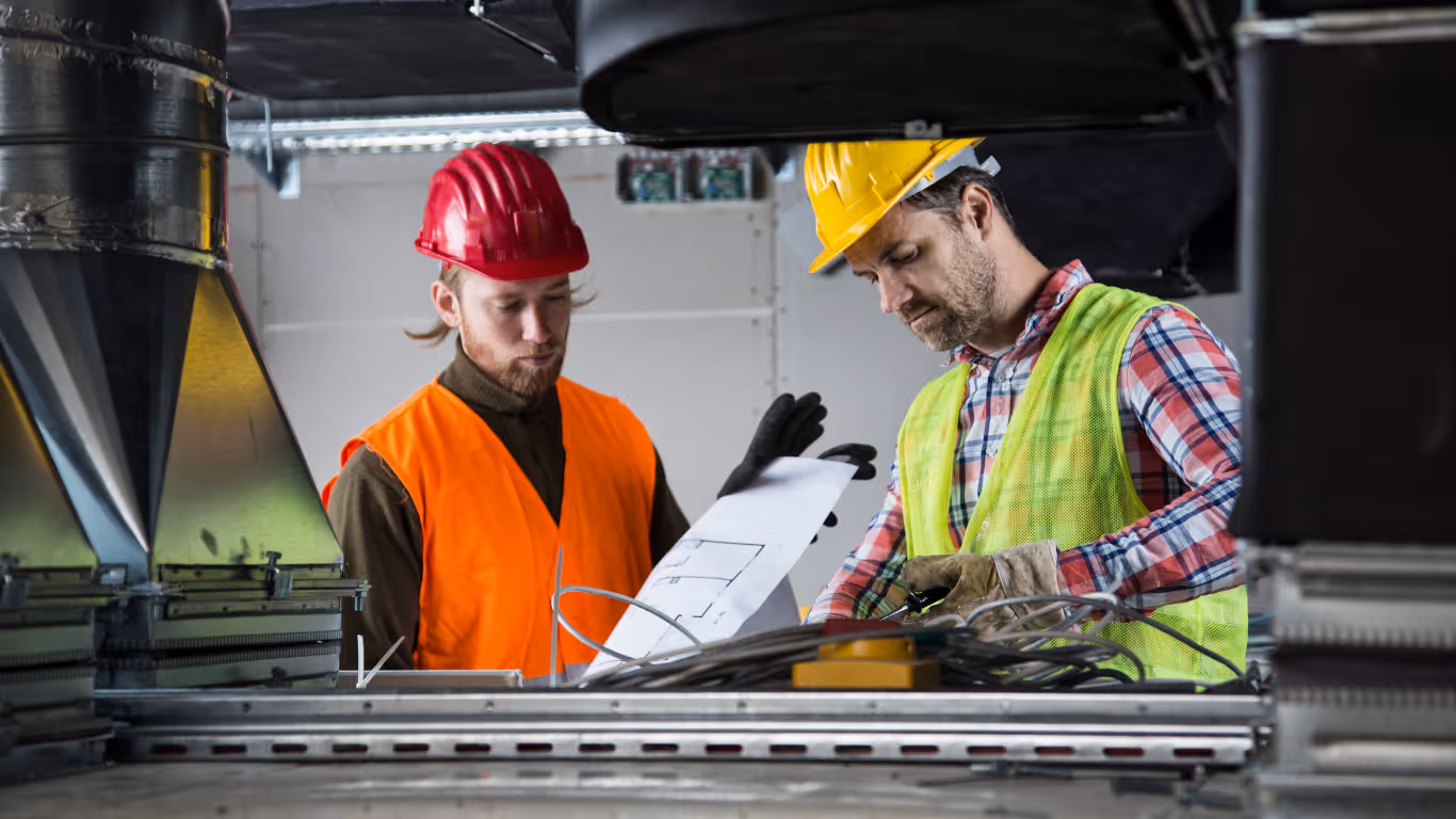 Two HVAC owners in hard hats and high-visibility vests review a blueprint together amidst industrial ventilation ducts and wiring.