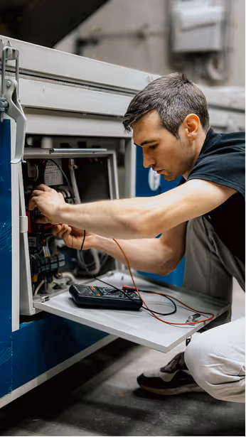 An HVAC tech in a black shirt kneels to examine electrical wiring inside an HVAC system with a multimeter placed on a shelf in front of them.