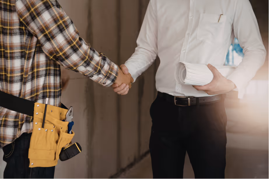 An hvac owner wearing a tool belt and a plaid shirt shakes hands with an investor in a white shirt holding rolled-up documents.