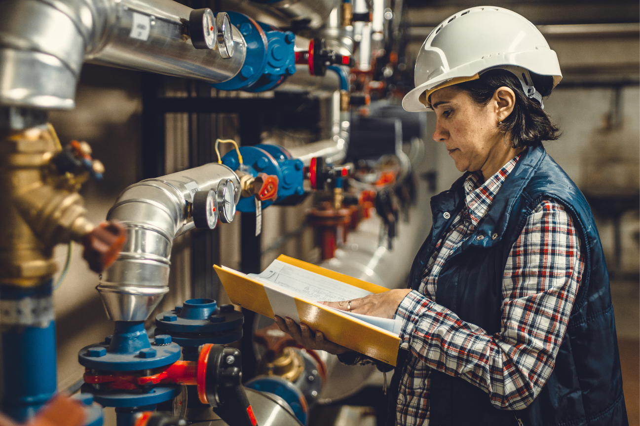 An hvac operator in a hard hat and vest examines a clipboard in front of complex industrial piping with gauges and valves.