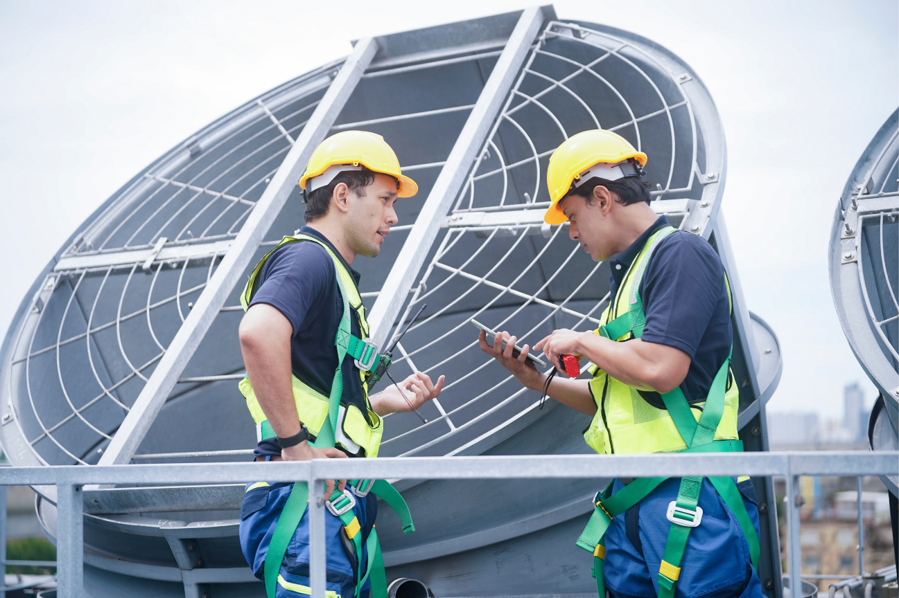 Two workers in yellow hard hats and high-visibility vests are standing on a rooftop, near large industrial cooling fans, discussing something on a tablet.
