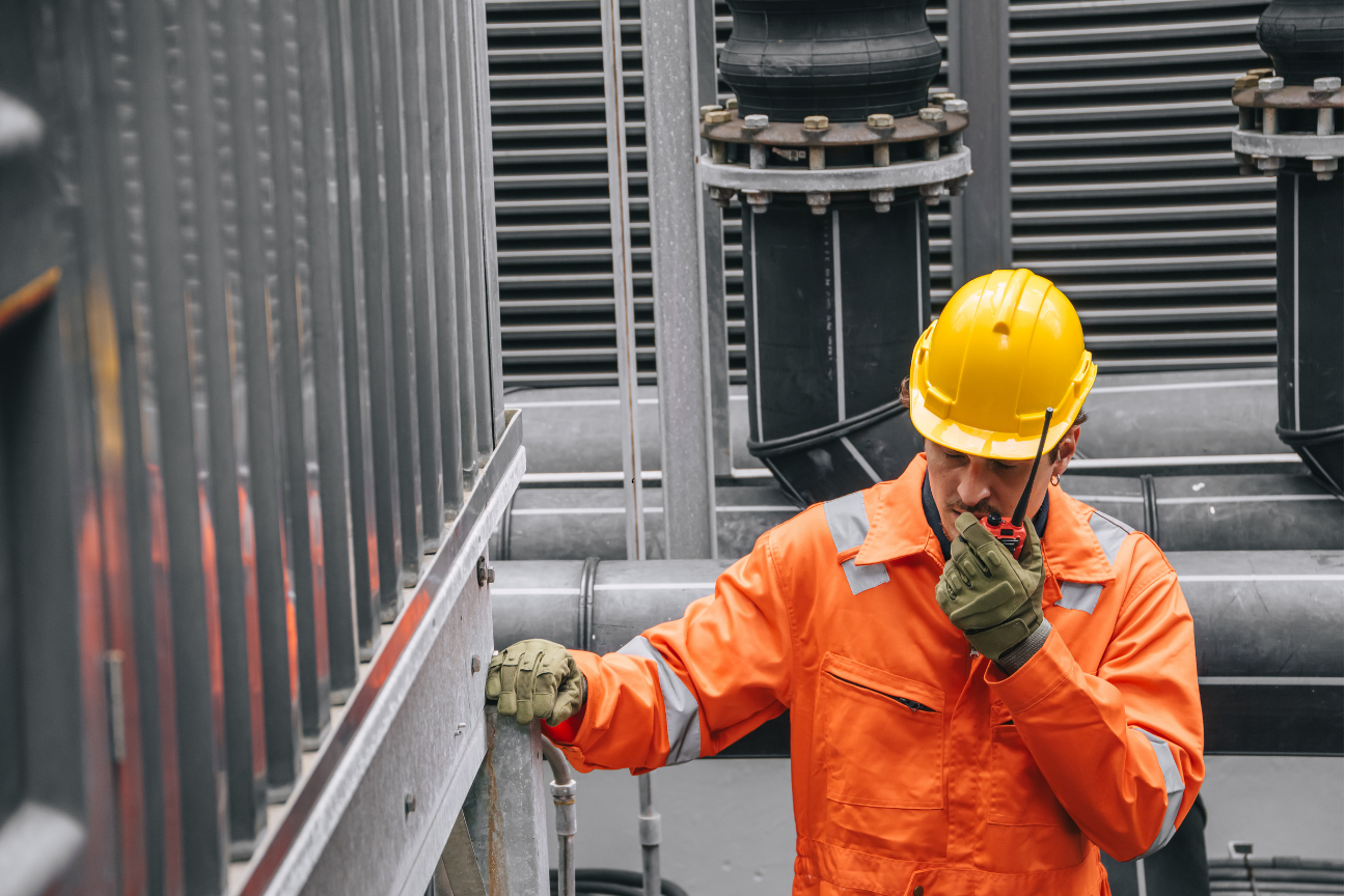 An HVAC tech wearing an orange jumpsuit and a yellow hard hat speaks into a walkie-talkie while standing next to industrial pipes and equipment.