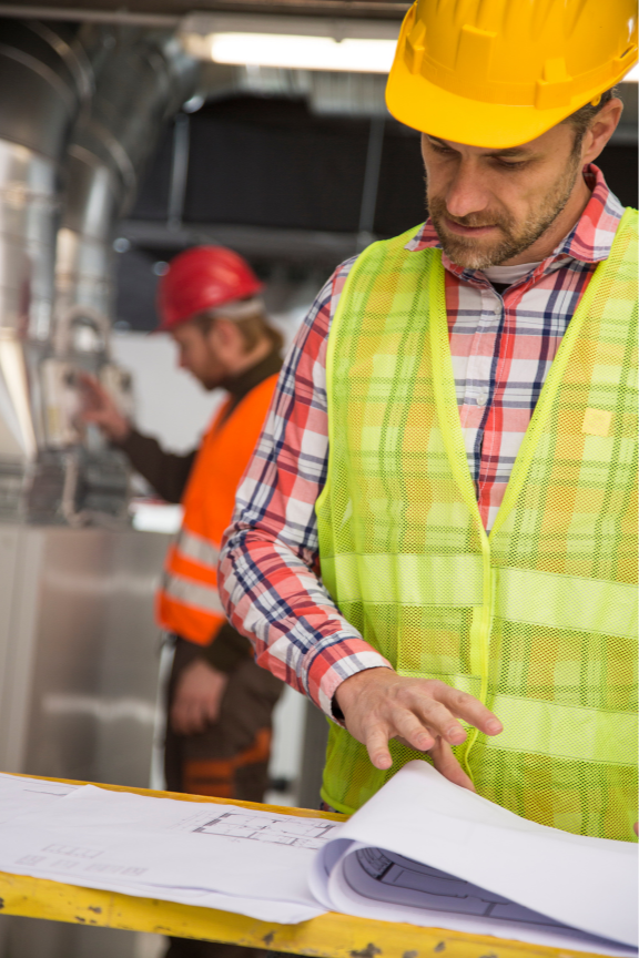 Two workers in hard hats and safety vests review hvac install blueprints on a table in an industrial setting.