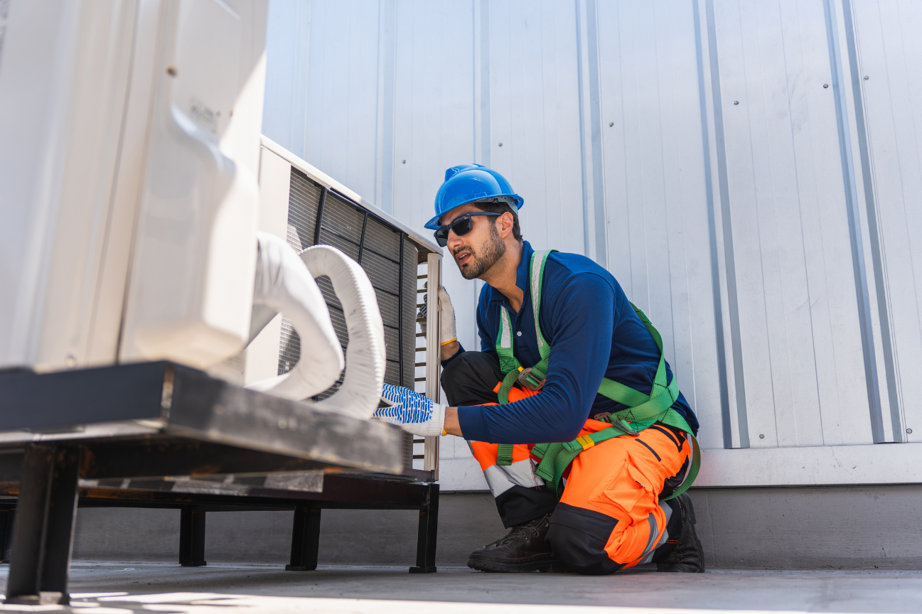 An HVAC tech wearing an orange jumpsuit and a blue hard hat services a rooftop unit.