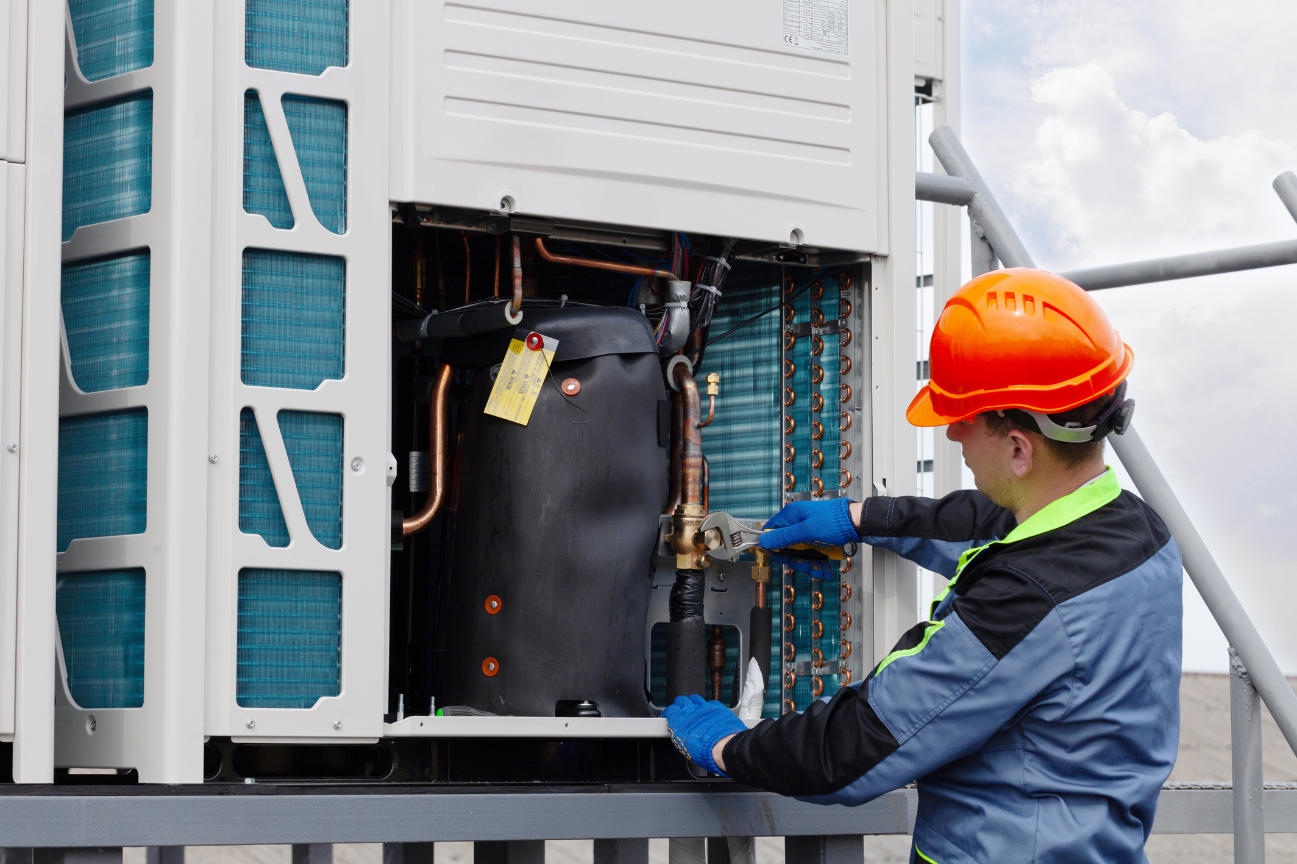 A worker in an orange hard hat and blue gloves repairs an outdoor air conditioning unit, using a wrench on copper pipes and connections.