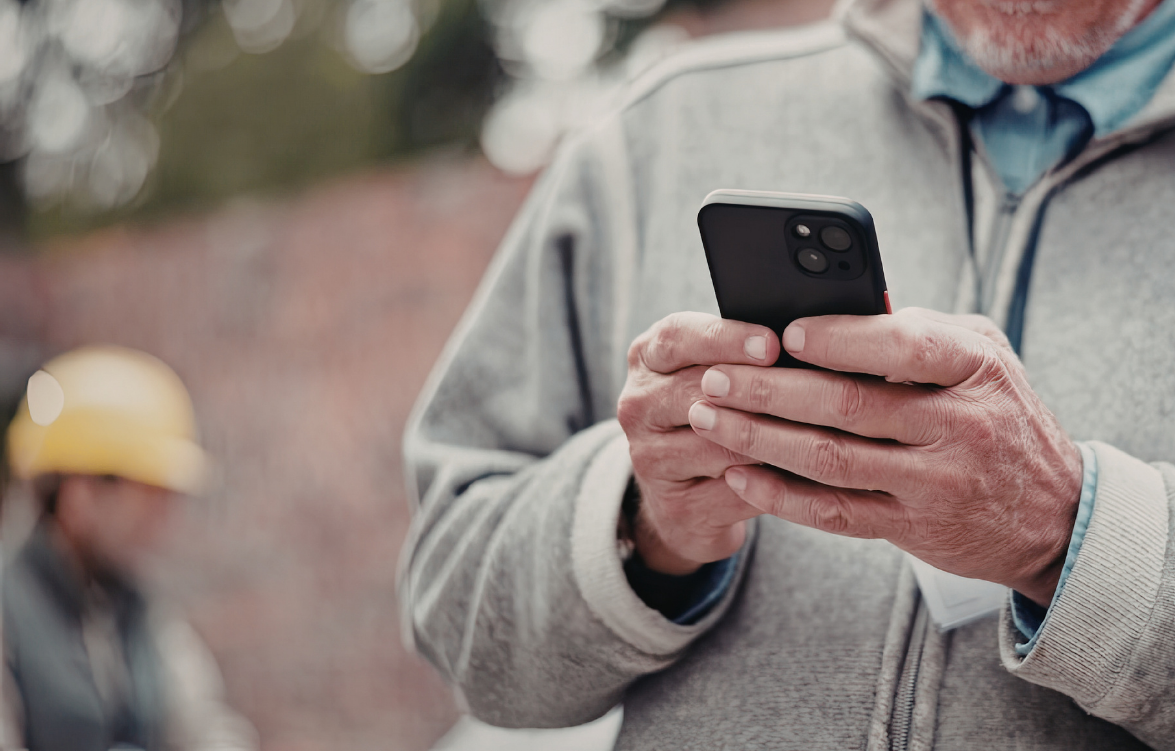 An HVAC in a grey jacket holds a black smartphone. In the blurred background, another person wears a yellow hard hat.