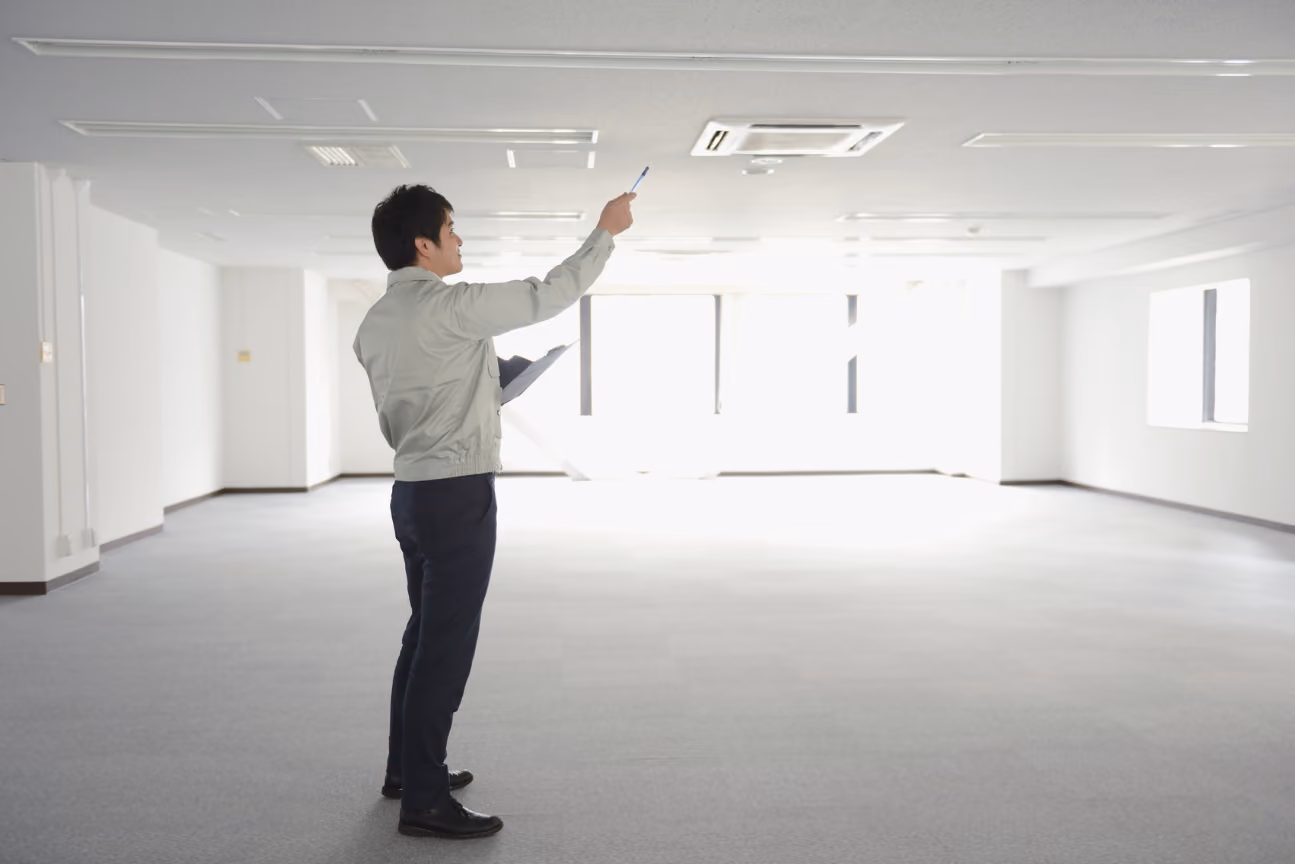 An HVAC tech in a light jacket and dark pants stands in an empty office space, holding a clipboard and pointing a pen towards the ceiling-mounted air conditioning unit.