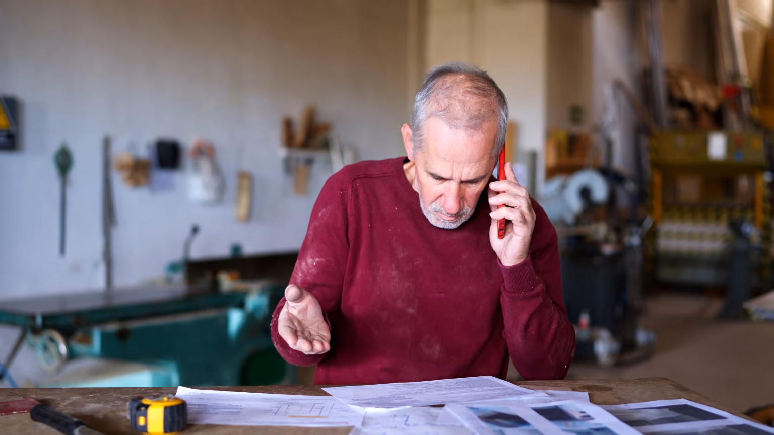 An HVAC owner in a workshop talks on a red phone while looking at papers on a workbench. Tools and machinery are visible in the background.