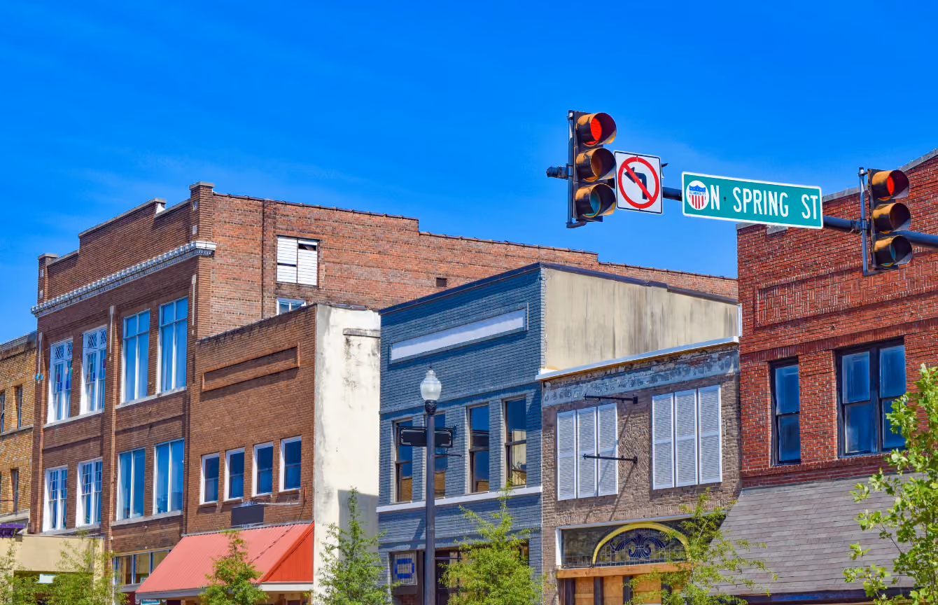Red brick and white commercial buildings under a clear blue sky, with a traffic light showing red and a street sign reading “SPRING ST” at an intersection.