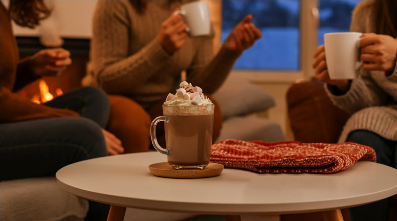 Glass mug of hot chocolate with whipped cream and marshmallows on a white table near friends by a fireplace.