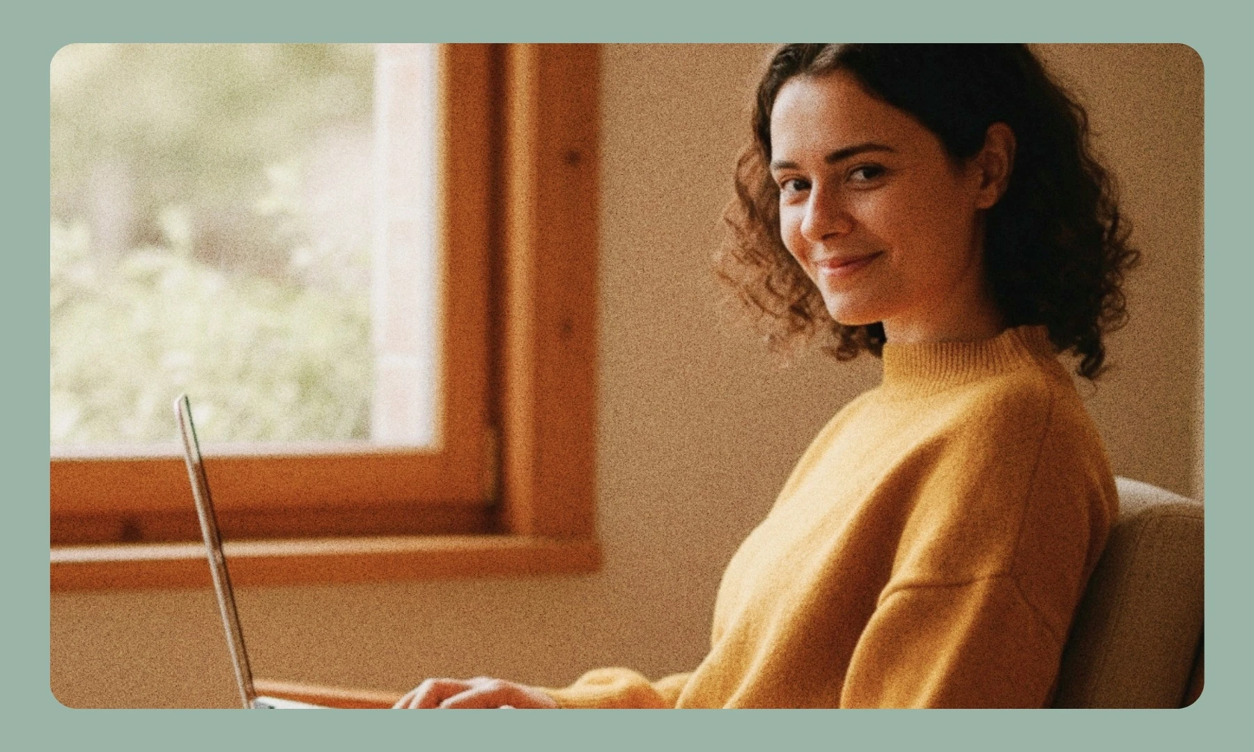 Smiling woman in a mustard jumper working on a laptop by a window