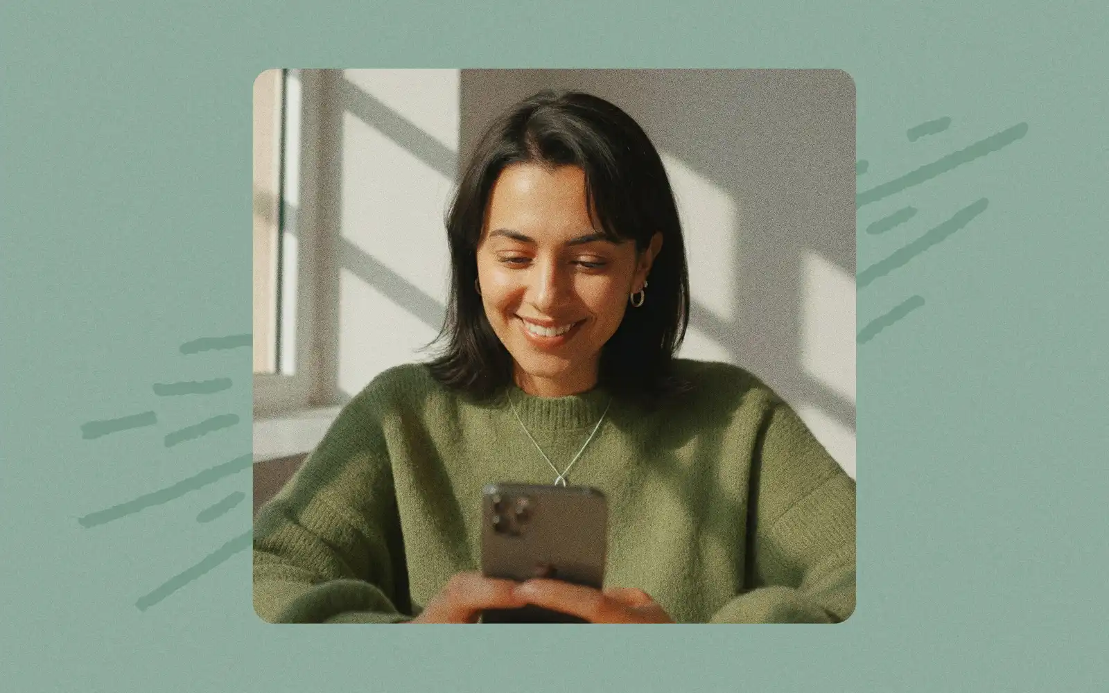 Smiling woman in a green jumper looking at her phone, set against a sage green background