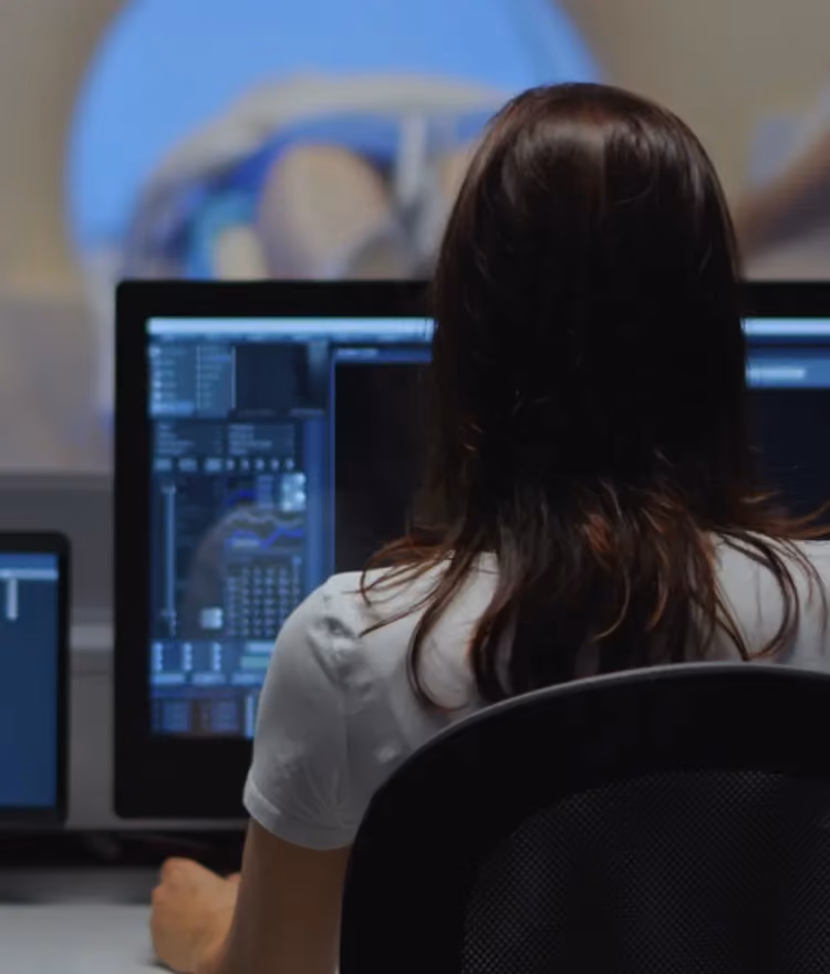 Medical technician monitoring diagnostic data on multiple computer screens while a patient undergoes an MRI or CT scan in the background
