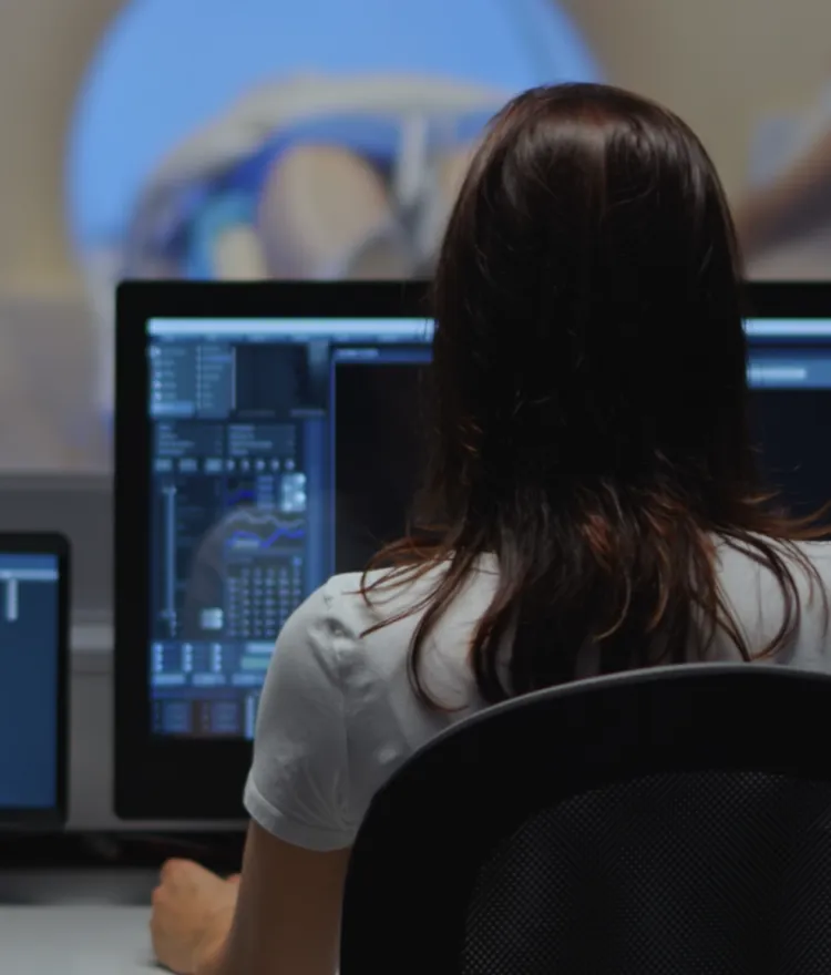 Medical technician monitoring diagnostic data on multiple computer screens while a patient undergoes an MRI or CT scan in the background