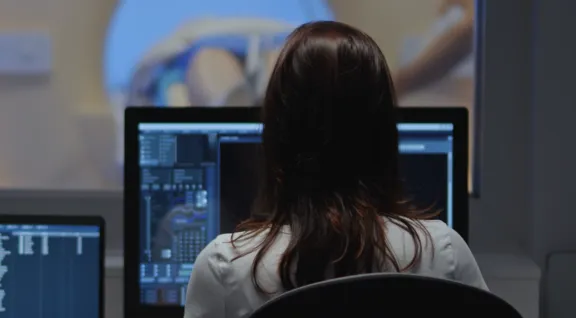 Medical technician monitoring diagnostic data on multiple computer screens while a patient undergoes an MRI or CT scan in the background