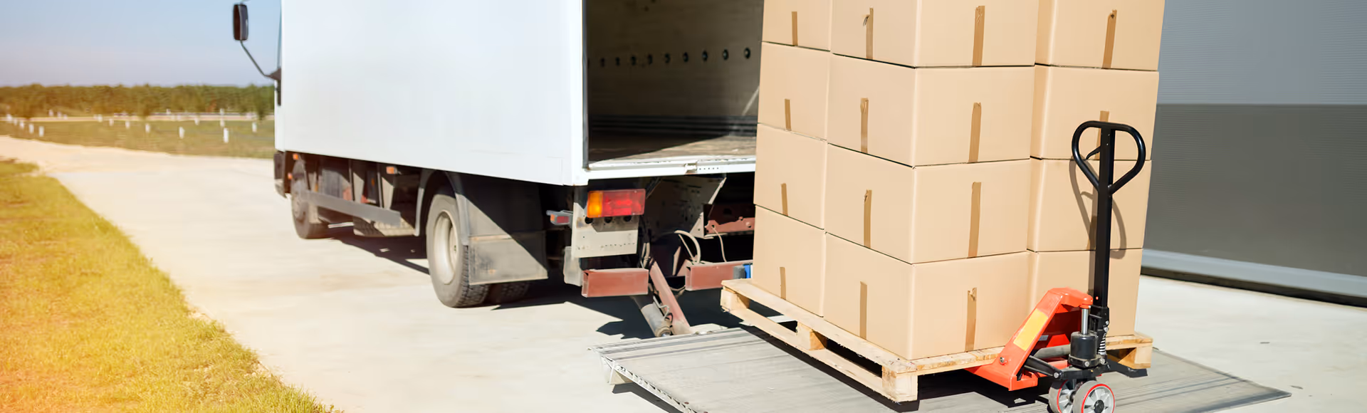 Pallet jack with stacked cardboard boxes being loaded into a white delivery truck on a sunny day