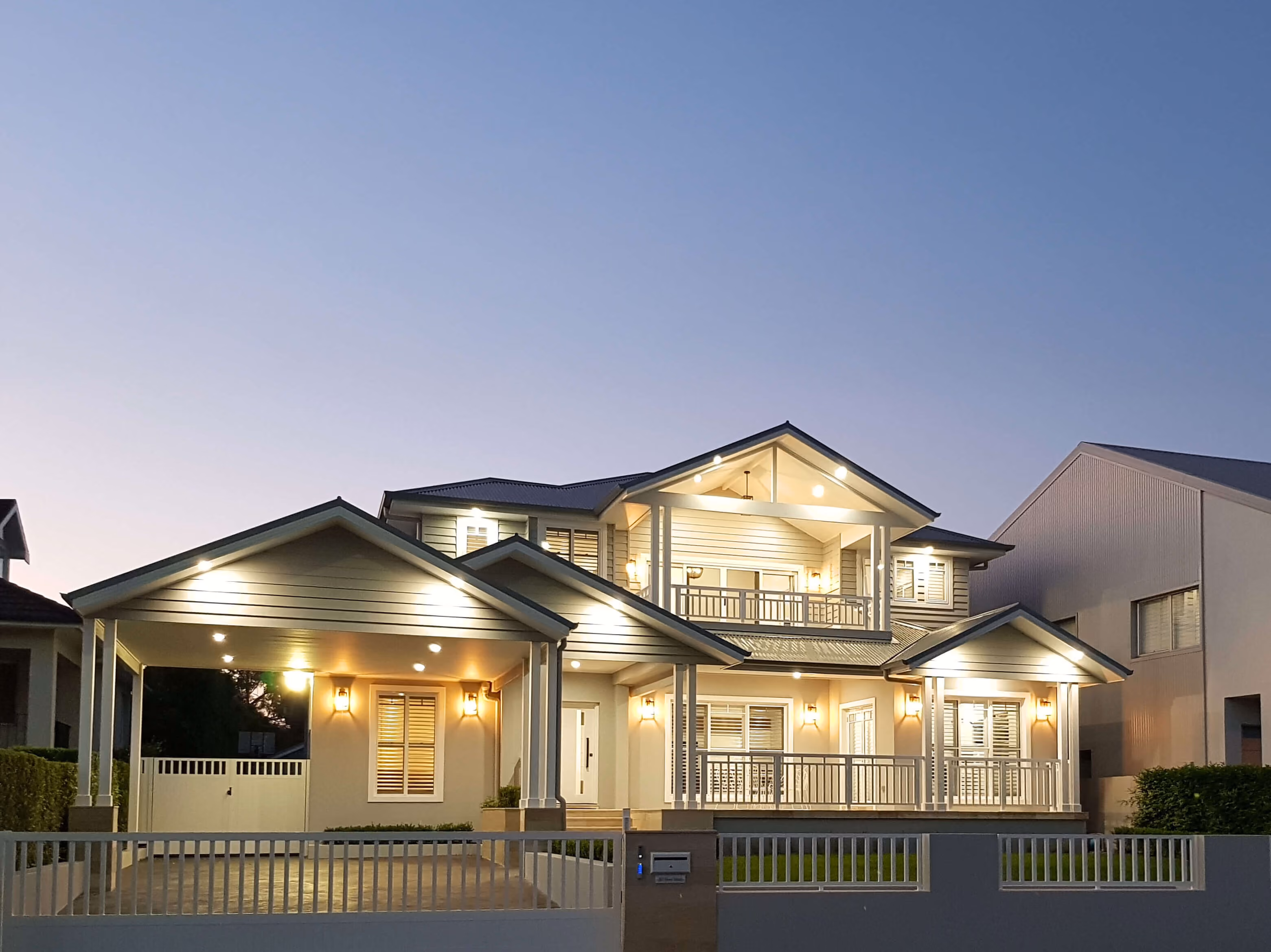 A two-storey Hamptons-style home photographed at dusk with warm interior lighting and a wide verandah.