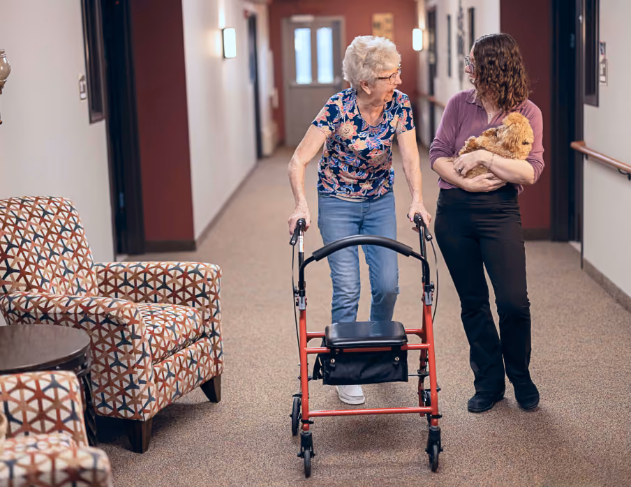 Elderly woman using walker and younger woman holding a small dog walking together in a hallway.