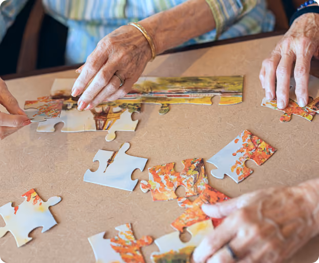 Elderly hands assembling a partially completed puzzle with autumn-themed pieces on a table.
